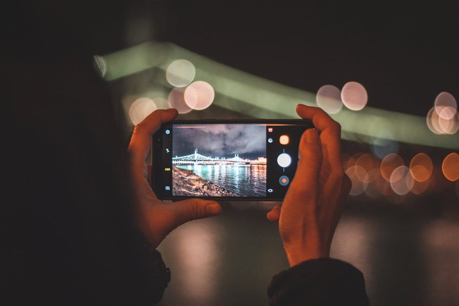 pexels-photo-4341198-4341198 Hands holding a smartphone capturing a cityscape at night with colorful bokeh lights.