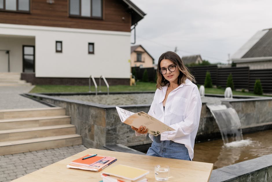 Woman in eyeglasses reading outdoors by a fountain with books on a wooden table.